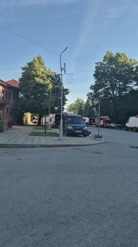 The video captures a street scene in Kirchevo, Bulgaria, on a clear, sunny day, likely morning or early afternoon. The camera initially points down a street lined with mature trees. A blue van, bearing the Bulgarian license plate "PB 9821 AC," is parked on the left side of the street next to a street lamp with a decorative banner. As the camera pans to the right, a market area unfolds with several stalls operating under red and white tents. Further down the street, various other vehicles, including a white van and a dark sedan, are parked. To the right, an outdoor cafe or seating area is visible with white umbrellas and several people gathered. Faint smoke or steam can be seen rising from the background near a cluster of trees. The reflection of the recording vehicle is visible in its side mirror on the right side of the frame. The scene depicts a calm, local market or village square with everyday activity.