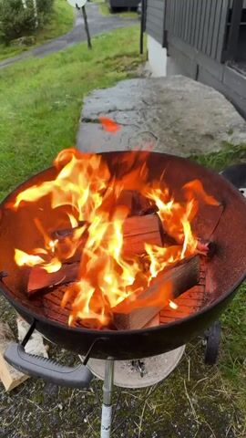 A close-up shot shows logs burning brightly in a charcoal grill. Flames leap from the wood, casting an orange and yellow glow. The grill, a classic black kettle style with handles and wheels, sits outdoors on a patch of grass. In the background, a stone path and a dark-colored building are visible, with greenery surrounding the scene. The camera is stationary.