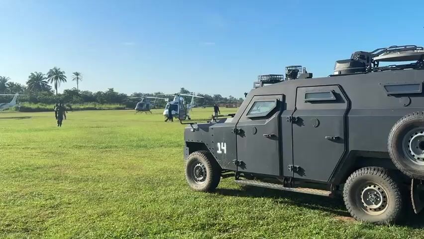 The video shows a grassy field with three helicopters parked and several people moving around. In the foreground is a gray armored vehicle with the number "14" painted on its side. The helicopters are white with green stripes and are facing in different directions. People are walking towards and around the helicopters. The sky is blue with a few clouds, and there are trees visible in the background. The overall setting appears to be a staging area or landing zone, possibly for security or transport operations.