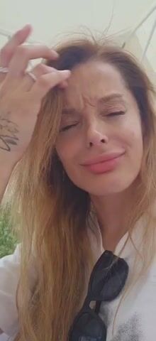 This is a close-up, first-person perspective video of a woman with long, blonde hair, wearing a white shirt. She is outside, likely under an umbrella, with greenery visible in the background. She touches her hair and makes various facial expressions, including smiles and pouts. A pair of black sunglasses is visible on her shirt. The video appears to be a selfie or a short, informal clip.