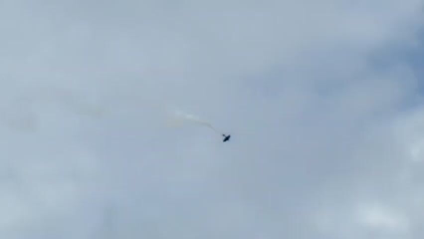 A pilot ejects from a jet that is trailing smoke. The pilot appears to be descending beneath the jet, attached by a cord. The sky is overcast.