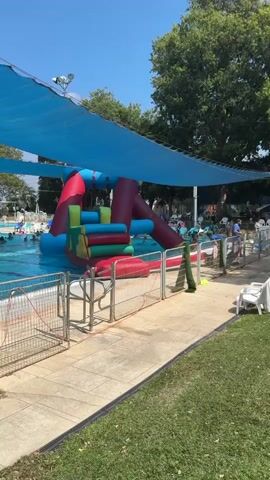 The video shows an inflatable water slide and obstacle course in a swimming pool. The slide is red, blue, yellow, and green. It is set up in the shallow end of the pool. There are people in the pool and around the pool. The area is shaded by a blue canopy. There is a grassy area to the right of the pool.