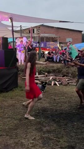 A woman in a red dress dances in a grassy outdoor area. A dog wearing a striped shirt and blue vest watches her. In the background, several people stand near a wooden structure with a banner. There are stacks of wood and debris scattered on the ground.
