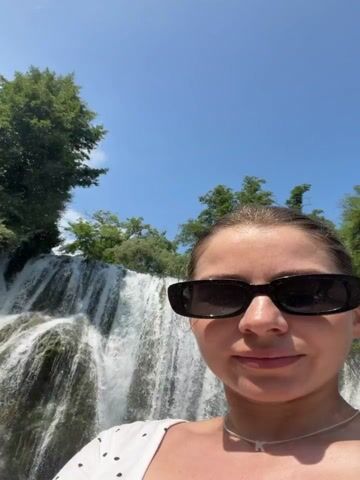 A young woman with blonde hair pulled back, wearing black sunglasses, a white top with black polka dots, and a delicate necklace, is taking a selfie in front of a beautiful waterfall. She is smiling and gesturing with her left hand towards the cascading water. The waterfall is wide and impressive, with water flowing down a rocky cliff face covered in greenery. The water collects in a clear, turquoise pool below. Lush green trees and foliage surround the top of the waterfall, and the sky above is a clear blue with a few scattered clouds, suggesting a bright, sunny day. The scene appears to be a natural tourist attraction, likely in a park or recreational area. The woman's expression is joyful and enthusiastic, indicating she is enjoying her visit. The context provided places this scene in Jajce, Bosnia and Herzegovina, a location known for its numerous waterfalls. There is no visible text in the media file.