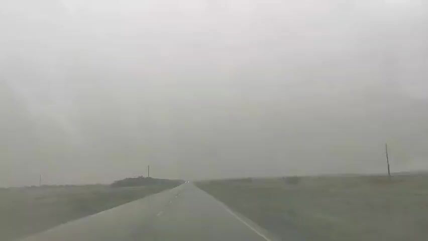 The video shows a first-person perspective of driving on a wet asphalt road under a heavily overcast and foggy sky. The road stretches into the distance, with green fields and scattered trees on either side. There are telephone poles visible along the side of the road. In the distance, a pair of headlights from another vehicle are faintly visible. The conditions suggest poor visibility due to the weather.