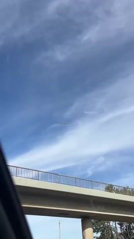A low-angle shot shows a concrete overpass with a railing against a blue sky with wispy clouds. A person is visible standing on the overpass near the railing. The camera then pans upwards to show a glider flying against the cloudy sky. The glider is shown from below, appearing as a silhouette against the clouds. The glider's wings are extended, and it is in flight. The video continues to show the glider moving across the sky.