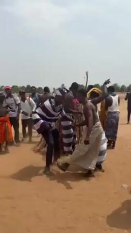The video shows a group of people gathered on a dry, dusty field. Several individuals are dancing, some are shirtless, and others are wearing traditional clothing with striped patterns. One person is covered in what appears to be straw. Other members of the group are dressed in more modern clothing like soccer jerseys or brightly colored suits. Dust is kicked up as the group dances in the middle of the field. The environment seems to be a rural area with trees visible in the background under a cloudy sky.