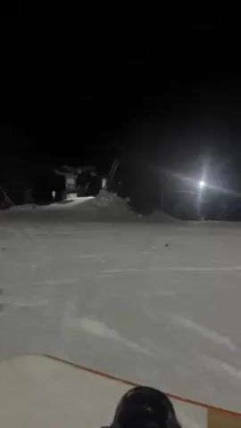 The video is taken from the perspective of someone standing on a snowy surface, likely a ski slope, at night. In the foreground, the edge of a snowboard is visible. The slope is illuminated by artificial lights. In the distance, a ski lift structure stands on the left, and a row of trees borders the slope on the right. Another person can be seen standing further down the slope, near the trees. It is a dark, snowy night scene.