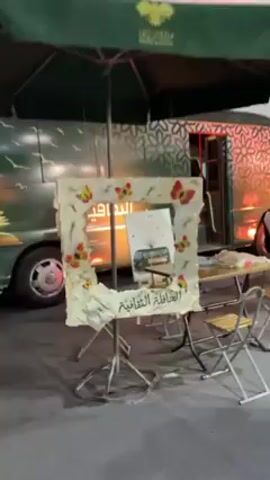 Outside near a bus, there's a decorated frame on a stand for taking photos. The frame is adorned with paper and butterfly embellishments. Arabic text is visible at the bottom. The bus in the background is painted with a pattern and has its lights on. Several people are moving around near tables and chairs. One man in a vest walks near the bus.