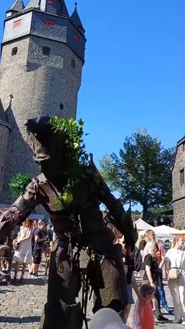 The video captures a lively outdoor event, possibly a festival or fair, set against the backdrop of a medieval-style stone castle with a prominent tower. In the foreground, a person in an elaborate Groot-like costume, made of what appears to be wood textures and green foliage, stands with one arm raised. A green apple is attached to the costume's chest area. Behind the Groot character, a crowd of people mill about, some appearing to be vendors with stalls and tents visible. To the right, another costumed performer, this one resembling a dragon with large wings and elaborate dark attire, stands on stilts. The ground is paved with cobblestones. The sky is a clear blue. The general atmosphere suggests a sunny day with a festive gathering.