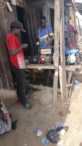 The video shows a man in a red shirt and black cap, standing near a table, and working on what appears to be a small engine. The man is wearing black pants and green sandals. He's focused on the object in his hands. Behind him, on the table, there are two portable generators, one red and one blue. Another man, wearing a blue patterned shirt, stands at the table, possibly working on the generators as well. The setting appears to be an outdoor workshop or repair area with metal sheets forming a wall behind the table. The ground is littered with trash and small debris.