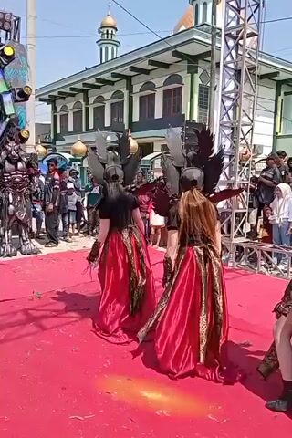 This is a short, vertical video shot outdoors during the day, likely a festival or performance. The foreground features a bright red carpet leading towards a stage area with metallic scaffolding. On the carpet, three individuals are dancing or performing. They are dressed in elaborate costumes, consisting of black tops, black shorts or skirts, and flowing, floor-length red skirts with gold trim. Each performer wears an intricate, dark, winged headdress that resembles a dragon or bird. Their movements suggest they are part of a synchronized performance. Behind the performers, a crowd of spectators stands watching, many dressed in casual or traditional clothing. To the left and right of the stage, more people are visible. In the background, a building with green trim, white walls, and multiple domes, resembling a mosque, is prominent. The sky is clear and blue. The overall atmosphere appears festive and engaging.