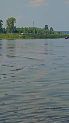 A boat with an orange and yellow stripe is moving fast on a wide river, kicking up a spray of water. The boat is moving from left to right across the frame. In the background, there is a green, tree-lined shore with a few buildings and a tall, thin tower visible. The sky is cloudy.