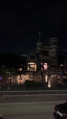 The video shows a building at night, likely a large house or mansion, with a tower-like structure on the right and a cross on top of a smaller turret. The building is illuminated from within, with some lights appearing pinkish. There's a fence in front of the building and a road. Cars pass by on the road. The sky is dark, and there are some trees surrounding the property. A car is parked on the right side of the frame.