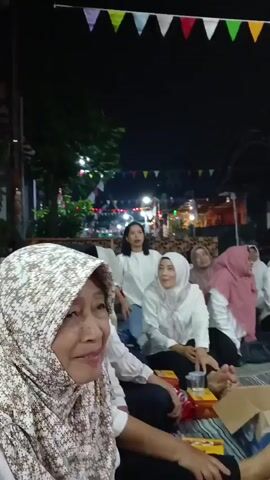 The video shows a group of people sitting on the ground at night, possibly at an outdoor gathering. There are colorful flags strung across the background, and some of the people are wearing white shirts. One person in the foreground is smiling and looking at the camera. The mood appears to be festive and relaxed.