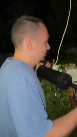 A person wearing a light-colored, ribbed t-shirt is seen from behind, actively operating a hand-pumped air blower with a black flexible hose. The hose is directed into a metal brazier, where a substantial fire of burning wood produces large flames and numerous glowing sparks. The person repeatedly pumps the device, causing the flames to intensify. The scene is set outdoors at night, with ambient lighting from string lights visible in the background, along with trees and a building. A second person in a light blue t-shirt is partially visible in the initial frames, observing the activity.