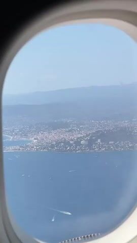 The video shows a view from an airplane window looking out at a coastal city. The city appears to be built on rolling hills and is situated along a body of water, likely the sea. Several boats are visible on the water. The sky is clear and blue, with some distant hazy mountains. The interior of the airplane, specifically the window frame, is visible in the foreground. The video is static, showing a single, unmoving perspective.
