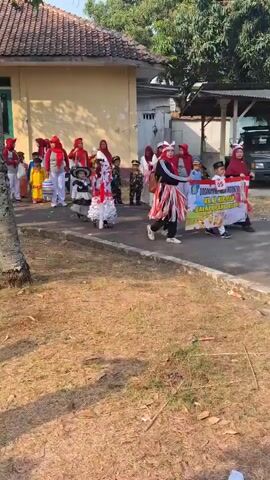 The video shows a group of children and adults gathered outdoors. Some children are wearing colorful costumes and are participating in what appears to be a parade or performance. One child in the foreground is wearing a white and red outfit with white ruffles and is being escorted by an adult in a matching outfit. Another child is wearing a blue costume with silver accents. Adults in the background are dressed in various casual clothing, with some wearing red headscarves. The environment appears to be a schoolyard or community area, with a building with a red tiled roof and a yellow facade in the background. There are trees and dry grass on the ground. A banner with Indonesian text is visible, suggesting a local event. Several people are holding cameras and phones, seemingly documenting the event. The overall atmosphere seems festive and communal.
