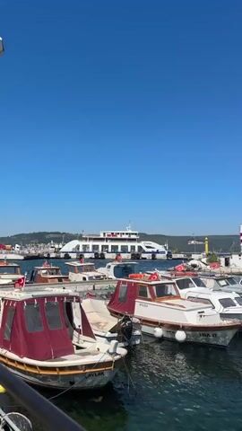This media file captures a bright, sunny day at a bustling harbor in Çanakkale Merkez, Turkey. The scene is dominated by a clear, vibrant blue sky and the calm, reflective waters of the harbor, dotted with numerous boats. The foreground features several smaller motorboats docked closely together, their hulls painted in various colors, some with awnings providing shade. Visible text on some of the boats includes "JDA 17," "ECESLI," "SIMGE 17," "HAYATIM II," and "B.K. 121707."

In the middle ground, a larger ferry is visible, suggesting active transportation and commerce. Further back, the shoreline is lined with more docked boats, a pier, and a lighthouse, its red and white stripes standing out against the blue sky. Beyond the harbor, the city of Çanakkale unfolds, with buildings and hills creating a picturesque backdrop. A tree stands tall on the left side of the frame, and a few people are walking along the promenade on the right, indicating a lively atmosphere. The overall impression is one of a picturesque, active port on a beautiful day.