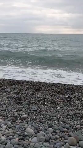 A choppy, gray ocean rolls onto a pebble beach under an overcast sky. A small wave breaks on the shore. A person swims in the distance on the right side of the frame. The camera is positioned on the beach, looking out at the ocean.