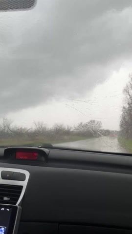 The video provides a first-person perspective from inside a car driving on a wet road during heavy rain. The sky is entirely overcast and grey, indicating poor weather. Numerous raindrops are visible on the windshield throughout the recording.

In the foreground, the interior of the car is visible, primarily the black dashboard with air vents. A red digital display mounted centrally on the dashboard shows "2.0 L/100," likely indicating fuel consumption, and "17:28," the current time. Below this display, an infotainment screen is visible, showing the "PEUGEOT" logo along with the time "16:29" and the date "03/07." A rearview mirror is also partially visible at the top left of the frame.

As the video progresses, the windshield wipers activate, sweeping across the glass to clear the rain. This action momentarily blurs the view before providing clearer glimpses of the road ahead. The road appears wet and is flanked by bare trees, suggesting a rural or semi-rural setting. In the distance, faint red lights, likely the taillights of another vehicle, can be seen. The continuous movement of the wipers and the persistent rain emphasize the challenging driving conditions under a gloomy, grey sky.