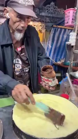 An elderly Asian man with a goatee prepares a crepe at a food stall. He wears a black jacket, a t-shirt with cartoon characters, and a baseball cap. He spreads Nutella from a jar onto a large, round crepe cooking on a griddle. His workspace is a small, cluttered food stall with various containers, a tip jar, and a colorful sign featuring a cartoon character.