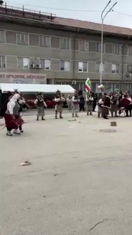The video shows a group of people dressed in traditional Bulgarian kukeri costumes performing a ritual in a town square. The kukeri are wearing ornate masks and animal skins, and they are carrying bells and other noise-making instruments. They are dancing and making loud noises to scare away evil spirits. A man in a black suit and brown shoes kicks a wooden block into the air. There are several people watching the performance, including a group of people standing near a tent and some other people standing near the building in the background. The sky is cloudy, and the buildings are gray.
