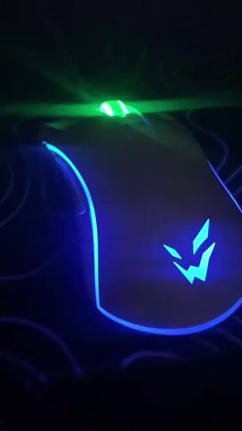 The video shows a gaming mouse with RGB lighting changing colors. The mouse has a white body with a logo on the base that lights up along with a strip around the bottom edge. The scroll wheel also has a green light. The background is dark with a mousepad visible with a pattern of intertwined lines. The color of the lighting changes from red to pink to purple.