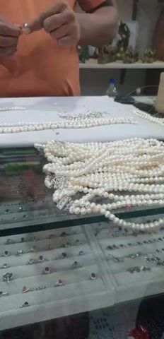 The media file shows a close-up, slightly blurry shot of a person's hands working on a pearl necklace. The hands are threading pearls onto a string. In the foreground, several strands of pearl necklaces are displayed, some with elaborate clasps. Below the necklaces, a display case filled with rings is visible. The setting appears to be a jewelry store or workshop. The lighting suggests it is daytime. The person is wearing an orange shirt. The background is out of focus but hints at shelves with items, possibly more jewelry or decorative objects. There is no visible text in the media file. The activity clearly depicts the crafting or assembling of jewelry. The overall impression is one of detailed work and craftsmanship.