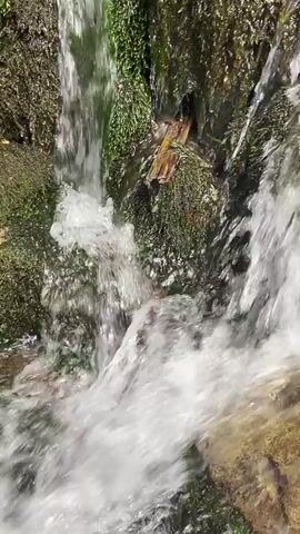 The video shows a close-up of a small waterfall cascading over moss-covered rocks. The water is clear and appears to be flowing rapidly. The rocks are covered in lush green moss, and there is a small piece of wood lodged in the moss. The lighting suggests it is daytime, possibly with sunlight filtering through trees. The camera is positioned close to the water, giving a sense of being immersed in the scene. There are no people or animals visible in the video. The overall impression is one of a natural and serene environment.