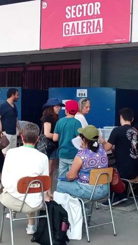 The video shows a group of people outside what appears to be a voting location. There is a sign above them that says "SECTOR GALERIA". The people are standing, sitting, and moving around. Some of them are looking at their phones or papers. There are chairs, tables, and voting booths visible in the background. The atmosphere seems calm and orderly. A woman sitting on a chair in the foreground looks at the camera briefly.