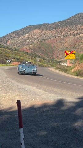 A blue Porsche convertible with two people inside drives around a curve on a paved road in a mountainous region on a sunny day. The car is the main subject, moving from right to left in the frame.  A yellow and red road sign in Arabic is visible on the right side of the road, along with a white and black chevron sign on the left. A red and white pole is in the foreground on the left.  The background features a clear blue sky and a large mountain with varied terrain.