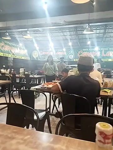 The video shows the interior of a restaurant. There are several tables and chairs set up. In the foreground, a bottle with a red and yellow cap is visible on a table. In the background, there are people sitting at tables and walking around. One person, a woman wearing a light-colored shirt, is standing and appears to be talking to someone. A banner with green text is hanging on the wall. The overall impression is a busy dining environment.