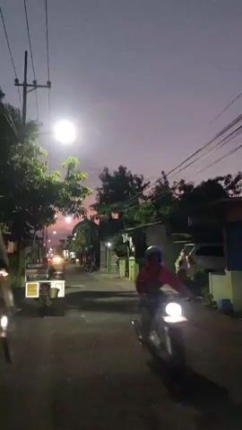 The video shows a street scene at dusk. A bright light, possibly a streetlamp, illuminates the foreground, casting shadows on the asphalt road. Trees line the street, their leaves dark against the twilight sky, which is a gradient of purple and pink. In the distance, car headlights and streetlights create a warm glow. A few buildings are visible along the sides of the street, some with lights on. A white truck with a yellow sign is parked on the left side of the road, and a motorcycle is parked on the right. A person is sitting on the curb on the right side of the street. The overall atmosphere is quiet and serene.