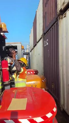 Two individuals wearing hard hats and safety vests are standing near a forklift. One individual, wearing an orange safety vest over a dark long-sleeved shirt and a black face mask, is holding a red fire extinguisher and appears to be attaching it to the forklift. The other individual, wearing a yellow safety vest over a dark shirt, is seated in the forklift. The forklift is red and grey, with white and red reflective tape on its side. A document is placed on the red hood of the forklift. The environment appears to be an outdoor industrial area with other forklifts and shipping containers visible in the background under a clear blue sky.