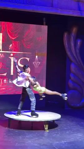 A male roller skater balances a female roller skater on his shoulders. The man wears sparkly pants and a vest. The woman wears a neon green leotard. They are on a circular stage with a white tablecloth. A large screen behind them displays the word "PARIS" in large letters. The background is dark, with some purple lighting.