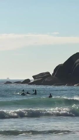 The video shows a group of surfers in the ocean near a rocky outcrop. The surfers are wearing wetsuits and are paddling on their surfboards in the turquoise water. Small waves are breaking in the foreground. In the background, large, dark gray rocks form the headland. The sky is blue with a few scattered clouds. A ship is visible on the horizon in the distance. The overall scene depicts a sunny day at the beach with people enjoying surfing.