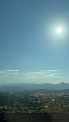 A vertical, handheld video displays a scenic view from a high vantage point. The sky is clear and bright blue with the sun prominently visible in the upper right quadrant. A few wispy clouds are scattered across the horizon. Below the sky, a range of rolling hills and mountains stretches across the landscape, appearing progressively lighter in color as they recede into the distance. In the foreground, there are patches of green foliage and trees, some of which are dark and dense, while others are lighter and more sparse. In the bottom portion of the frame, there is a railing or barrier, partially obscuring the view, and the ground appears to be covered in gravel or dirt. The video captures a sense of a vast, open outdoor environment under a sunny sky.