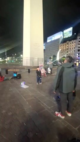 At night in a city square, a tall white obelisk is visible. A man wearing a gray and black jacket and red and white sneakers is standing on the cobblestone ground, facing the obelisk. He appears to be taking photos or videos with his phone. A small child in a dark jacket and jeans is standing near the base of the obelisk, looking towards the man. Other people can be seen in the background, some also holding up their phones. Behind the obelisk are several tall buildings illuminated by lights. A small audio system is placed near the base of the obelisk. The overall atmosphere seems festive or touristy.