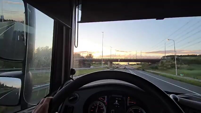 The video shows a first-person perspective from the driver's seat of a truck. The truck is on a highway. The sun is setting, casting a warm glow on the scene. The driver's hands are visible on the steering wheel. The dashboard of the truck is also visible, with various gauges and indicators. In the distance, there is an overpass and power lines. The sky is clear with some clouds. The overall impression is one of a long journey as the sun sets.