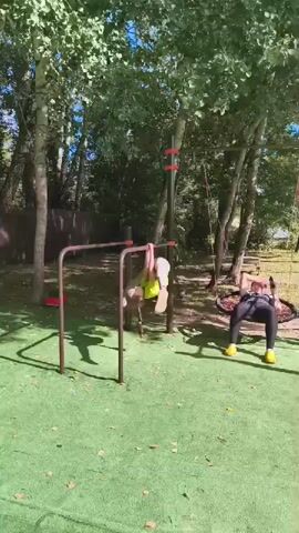 Two women are exercising outdoors on a playground. The woman on the left is performing a dynamic exercise on a horizontal bar, transitioning between hanging and leg splits. She is wearing a neon yellow top and gray leggings. The woman on the right is seated on a swing and appears to be doing some form of exercise, possibly core work, as she is bent forward with her hands on the ground. She is wearing black leggings and a black top. The environment is a park with artificial turf, trees, and playground equipment.