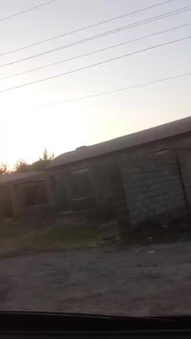 The video shows a view from inside a vehicle moving along a road. On the right side of the frame, there are several buildings and structures. The first building appears to be made of concrete blocks and has a curved roof. Adjacent to it is a larger building with a dark roof and a green awning. This building has a fence in front of it and what appears to be a sign with some writing on it. The interior of this building is visible through the fence and windows, showing multiple levels and windows. Further down the road, another building or structure can be seen. The environment appears to be somewhat industrial or rural, with some overgrowth and scattered debris visible. The sky is clear and bright, suggesting daytime. The video is captured from a moving vehicle, indicated by the slight blur and the presence of a side mirror.