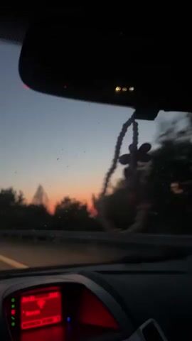The video is a first-person perspective from inside a car, looking out the windshield and at the rearview mirror. The time appears to be sunset, with a gradient of orange and purple in the sky. Trees and foliage line the road, which seems to be a highway. A beaded car accessory with a flower charm hangs from the rearview mirror and sways slightly. The car's dashboard displays a red screen with what appears to be radio information, showing a WiFi symbol and the text "Bana". The video is slightly shaky, indicating movement. There are no people visible.