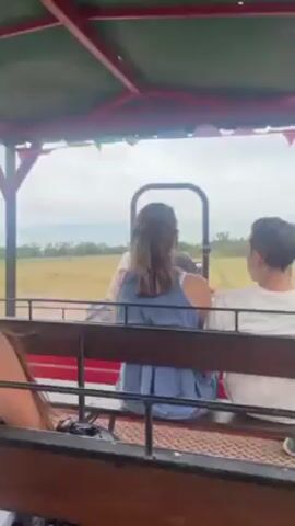 The video shows a group of people riding on a trailer-like vehicle. The trailer has a red frame, wooden benches, and a canopy adorned with colorful flags. The passengers are seated and appear to be enjoying the ride, with some conversing with each other. The camera is positioned from the perspective of one of the passengers, showing their lap, adorned with a tattoo, and a small bag. The landscape outside consists of a wide, open field with dry grass and scattered trees under a cloudy sky. The trailer is moving, as evidenced by the changing scenery.