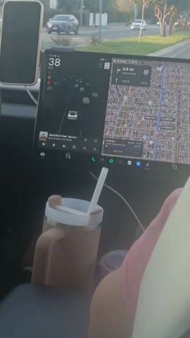 The camera is positioned inside a moving Tesla, capturing a passenger-side view. The car's dashboard features a large display screen showing navigation and music playback. A smartphone is mounted to the left of the screen. A beverage container with a straw rests on the passenger seat. The car is traveling on a city street, passing by other vehicles and street signs. The speedometer displays the car's speed, fluctuating between 36 and 38 mph. The environment appears to be a residential or suburban area with trees lining the streets.