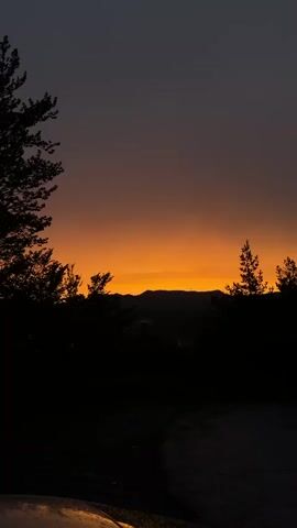 A silhouette of trees and mountains is visible against a vibrant orange and dark purple sunset sky. The foreground shows a dark area, possibly a road, with a faint reflection of light. The scene is captured from a low angle, looking upwards towards the sky and distant landscape.
