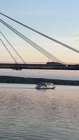 A wide shot of a cable-stayed bridge crosses a body of water. A large, white passenger boat with multiple decks travels on the water beneath the bridge. The boat is moving from left to right in the frame. The bridge has two tall, white and red striped pylons, from which cables extend to support the bridge deck. Vehicles are visible driving on the bridge. The sky above is a clear blue with some pale pink and orange hues near the horizon, suggesting either sunrise or sunset. A tree-lined shore is visible on the far side of the water. The water is calm, with gentle ripples. The foreground shows the edge of the water with some grass and gravel.