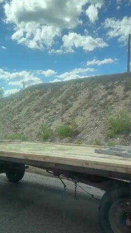 The video opens on a cloudy day, with a clear blue sky visible. The camera is positioned inside a vehicle, looking out at a rural scene. In the foreground, a flatbed trailer is visible, its wooden surface weathered and worn. The trailer is attached to a tractor, which is partially obscured. The tractor is pulling a green implement with the word "MARKANT 50" visible in red lettering. The implement appears to be some sort of agricultural machinery. The tractor and implement are moving along a paved road. In the background, a dry, rocky hillside is visible, dotted with sparse vegetation. Further in the distance, a tall, slender structure, possibly a smokestack or tower, can be seen against the sky. The camera continues to move, capturing the passing landscape. The overall impression is one of agricultural activity in a somewhat arid environment.