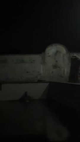 A dark, outdoor environment is depicted at night, likely a courtyard or patio. The ground appears to be wet or reflective, suggesting recent rain or a water feature. In the middle ground, a wall with peeling paint and some architectural details, possibly an alcove or arch, is visible. To the left, a small fountain or water spout is visible, with water gently falling into the dark pool below. The overall lighting is dim, with the sky completely black. There are no people or animals visible in the frame. The scene is quiet and still, with the primary focus being the interplay of light and shadow on the wet surfaces and the ancient-looking wall.