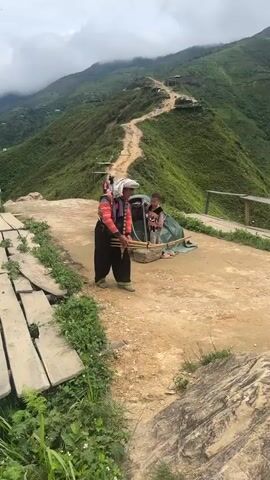 A video shows a person in traditional clothing playing a musical instrument in a mountainous area. The person, wearing a headscarf, a red and black jacket, and black pants with striped cuffs, is holding and playing a bamboo instrument that appears to be a panpipe or a similar multi-piped wind instrument. To the left of the person is a child in a green tent. In the background, a winding dirt path leads up a lush green mountain, with scattered structures visible along the path. The terrain is steep and covered in vegetation, with a wooden walkway in the foreground. The sky is overcast. The person is in motion, suggesting they are walking or performing.